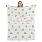 A woman is holding up a blanket with a repeating pattern of green frogs and the name "Francesca" written on it.
