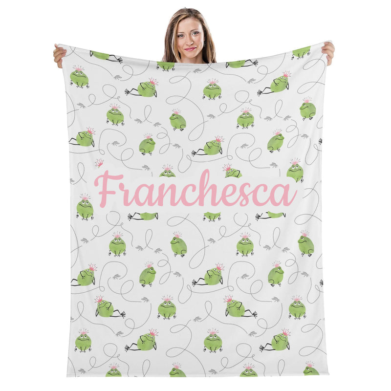 A woman is holding up a blanket with a repeating pattern of green frogs and the name "Francesca" written on it.