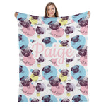 A woman is holding up a colorful blanket with a repeating pattern of pug illustrations and the name "Paige" written on it.