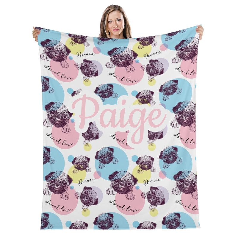 A woman is holding up a colorful blanket with a repeating pattern of pug illustrations and the name "Paige" written on it.