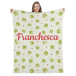 A woman is holding up a blanket with a repeating pattern of green frogs and red hearts.