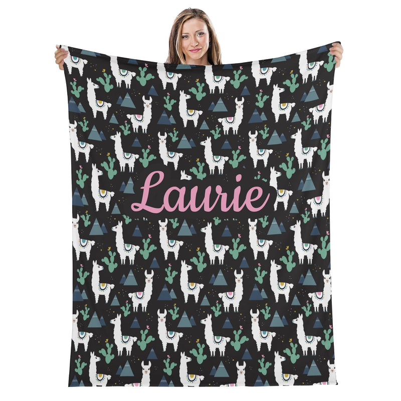 A woman is holding up a black blanket with a repeating pattern of white llamas and cacti on a black background.