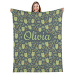 A woman is holding up a large blanket with a repeating pattern of green owls and leaves on a dark gray background.