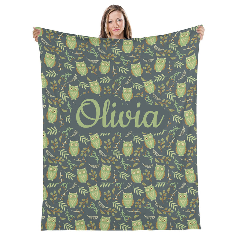 A woman is holding up a large blanket with a repeating pattern of green owls and leaves on a dark gray background.