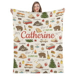 A woman is holding up a blanket with a repeating pattern of camping-related objects and activities, including a tent, RV, campfire, and various outdoor elements.